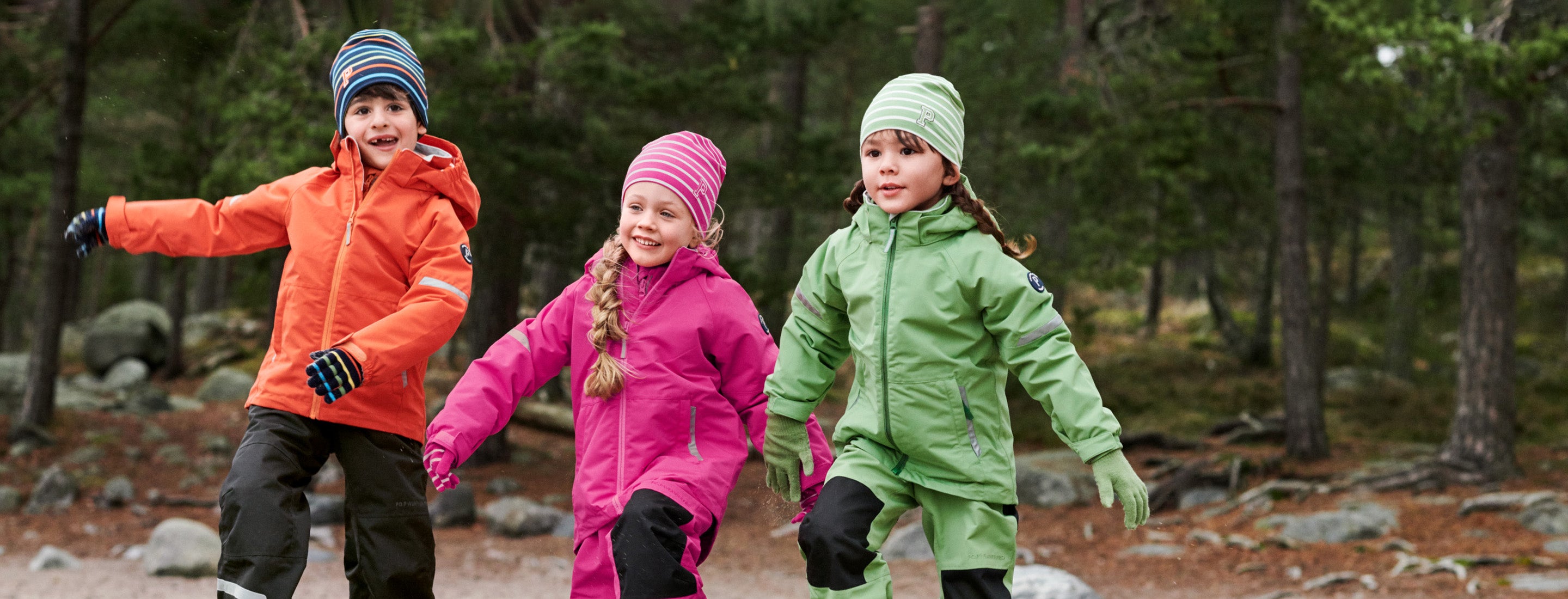 Three children in a forest wearing Polarn O. Pyret windbreakers and beanies  