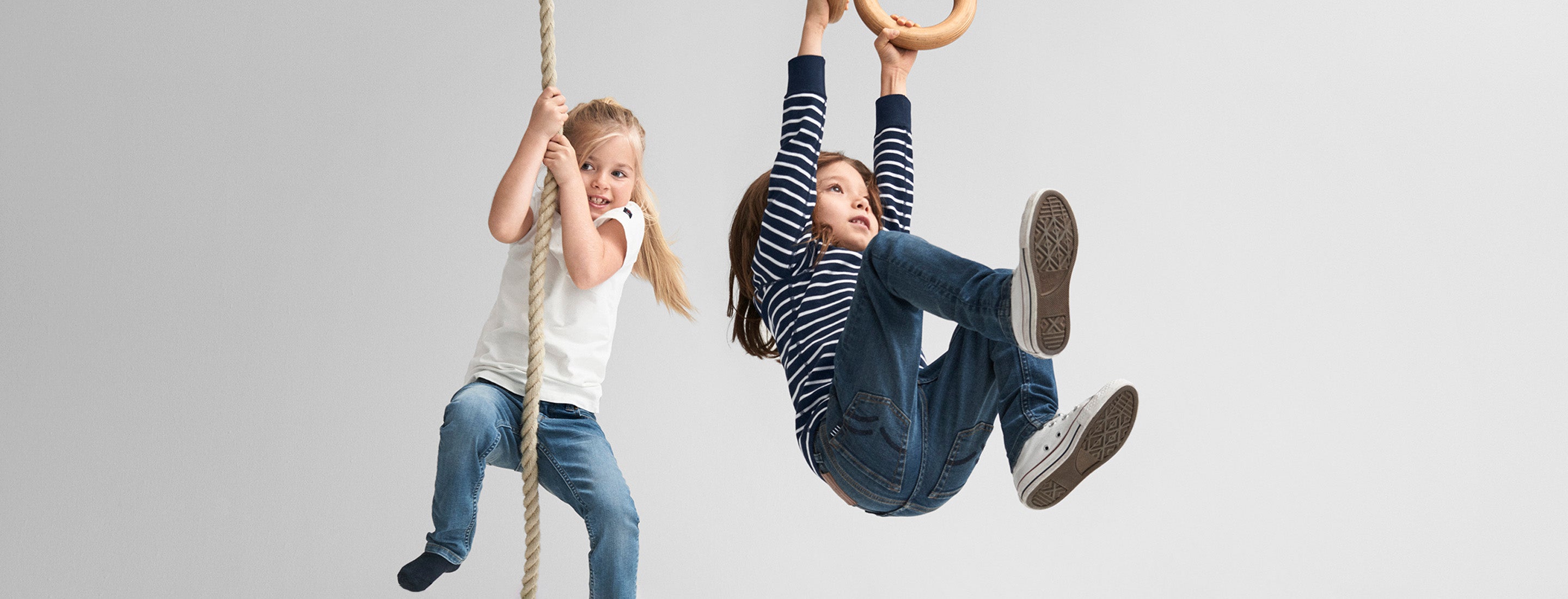 One child climbing a rope wearing a white Polarn O. Pyret shirt and Polarn O. Pyret jeans and one child climbing a gymnastic ring wearing a striped Polarn O. Pyret shirt and Polarn O. Pyret jeans 
